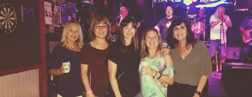 Five women pose with a band in a bar. One woman holds a drink, the band plays on a stage.
