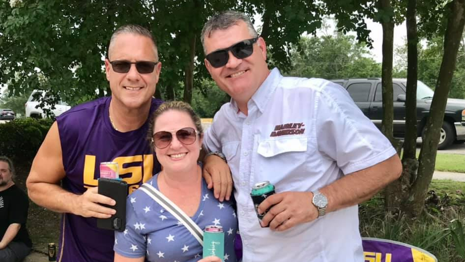 Three people smiling, posing outdoors. Two men wear sunglasses, one in LSU shirt. Woman in sunglasses, stars shirt, holds a drink.