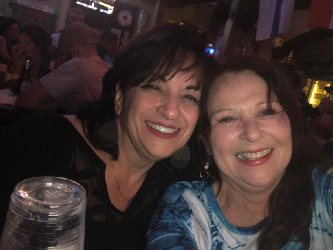 Two smiling women, close-up, indoors. One in black top, other in blue tie-dye. Bar setting, blurry background.