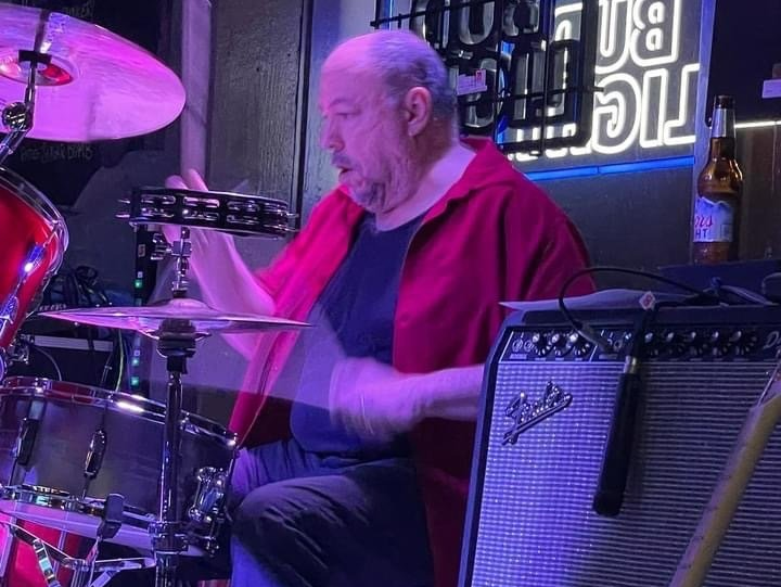 Drummer playing the drums and tambourine at a bar, wearing a red shirt, with neon sign in the background.