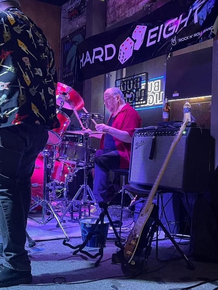 Drummer playing at Hard Eight bar; drums, amp, bass guitar, stage lights.