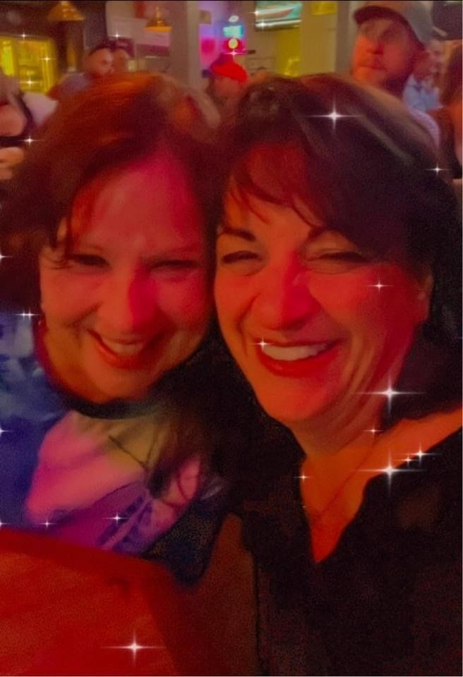Two smiling women pose close, illuminated in warm light, likely at a social gathering.