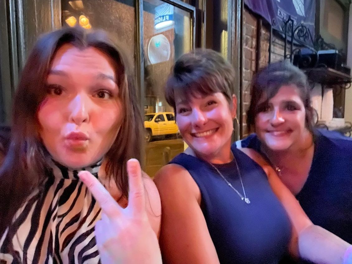 Three women smiling at the camera, posing outdoors at night. One makes a peace sign.
