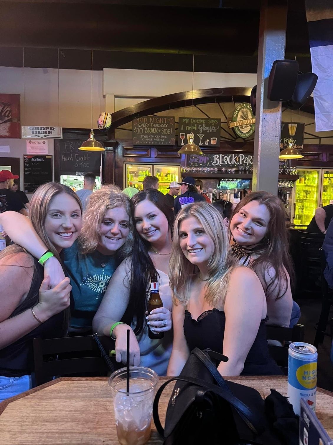 Five women smiling at a table in a bar; one holding a beer bottle, one with an arm around another.