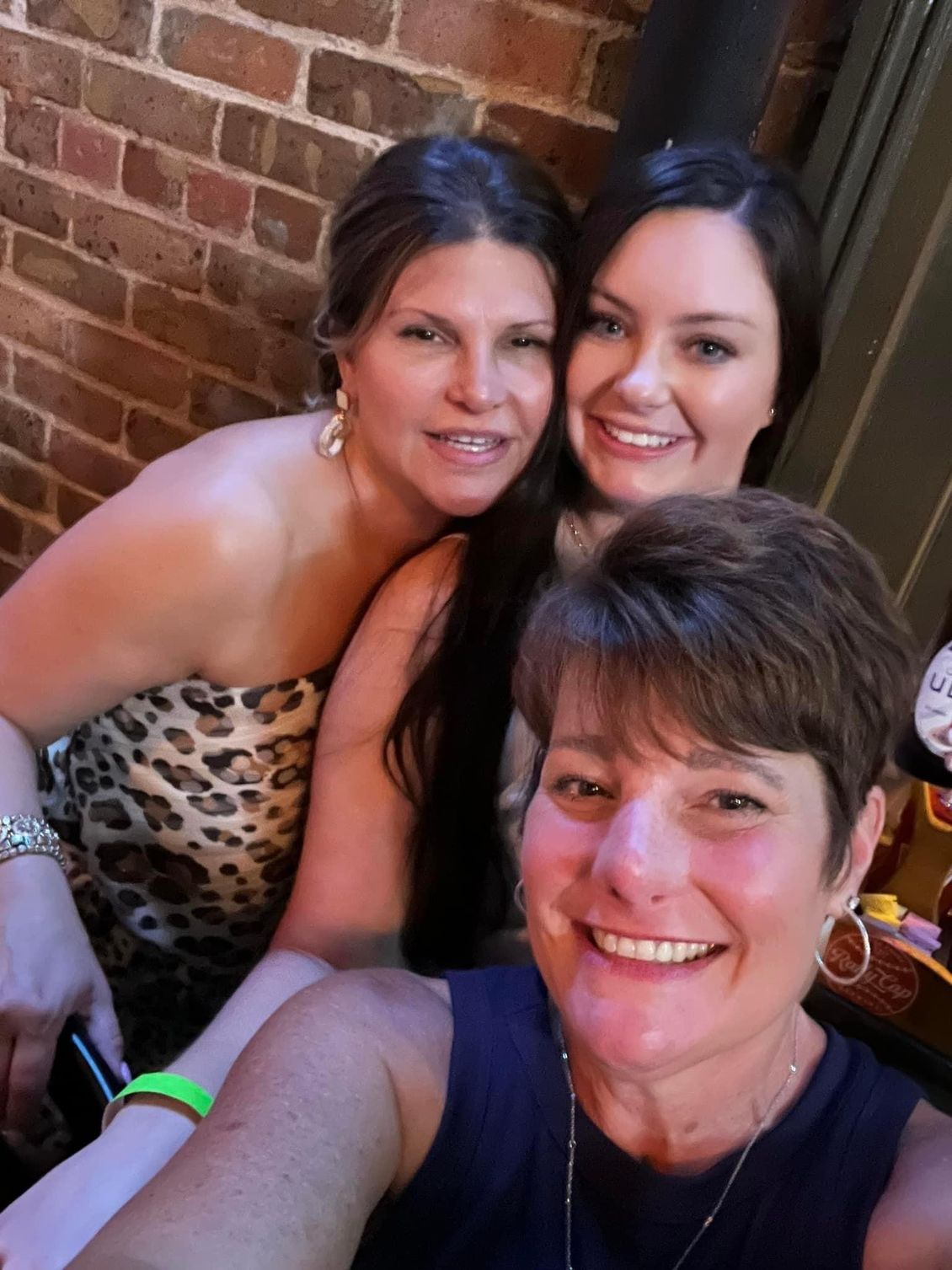 Three smiling women posing for a selfie in front of a brick wall. One wears a leopard-print top.