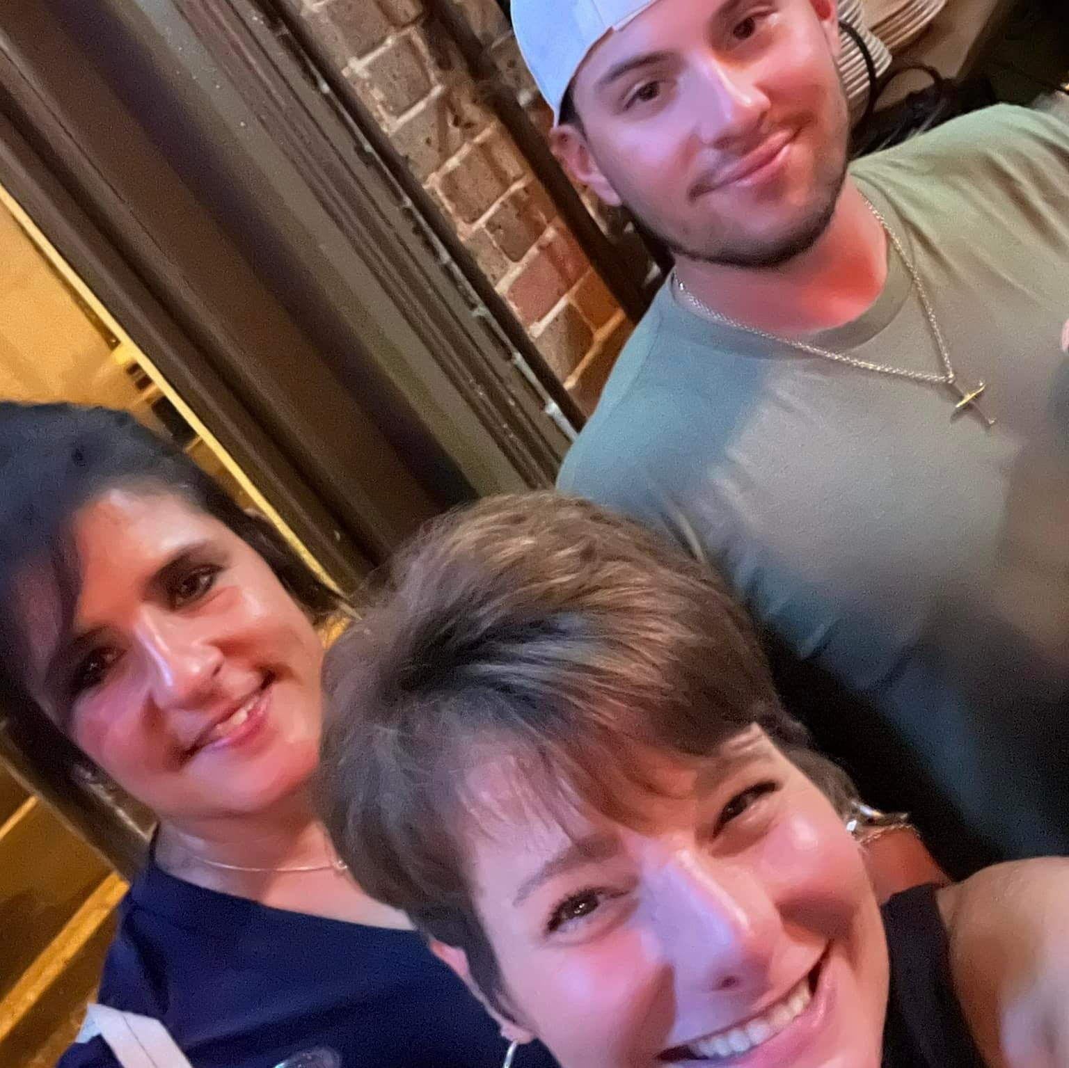 Three people smiling at a bar: two women in front, one man in the back. Brick wall visible.
