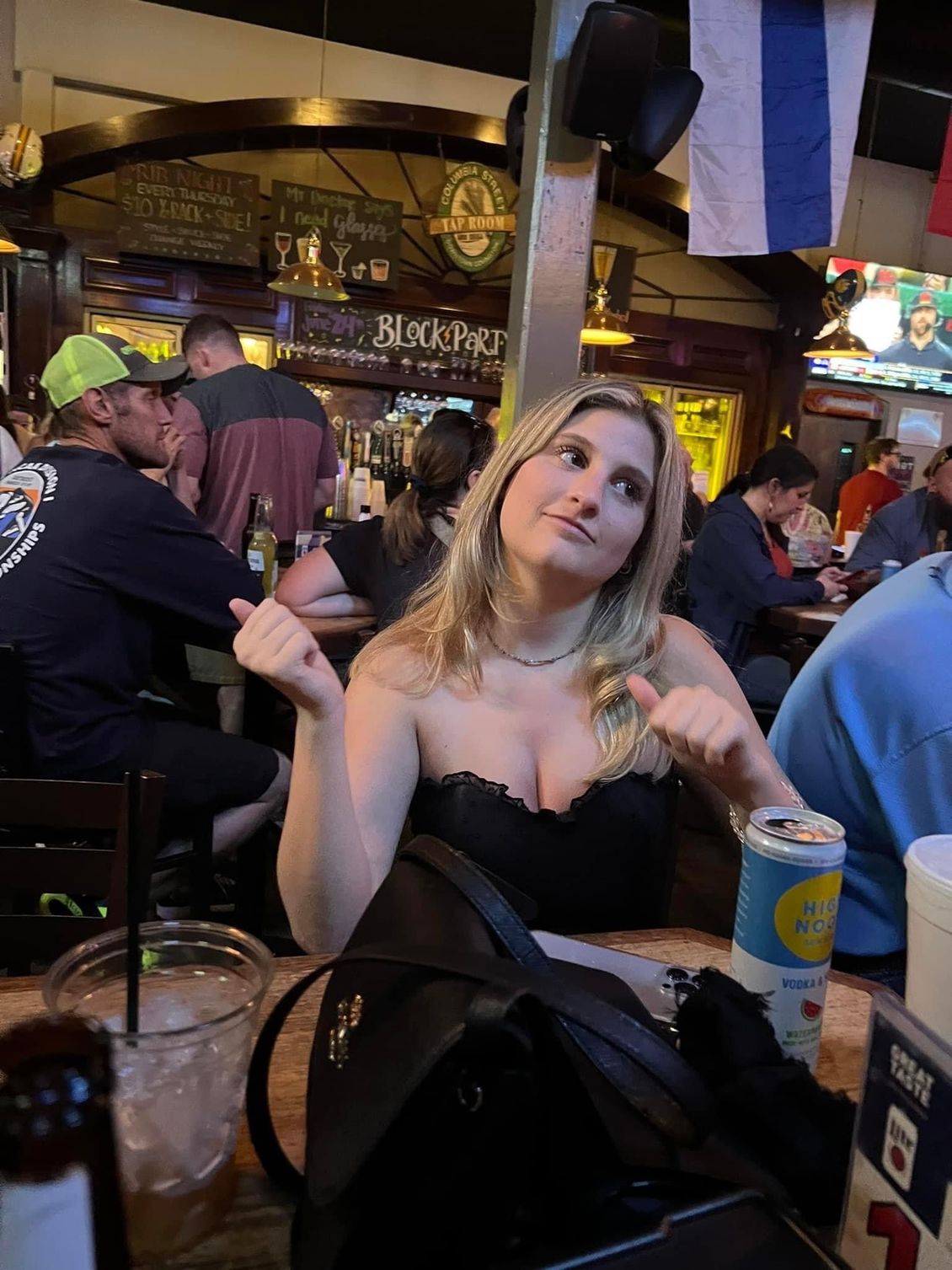 Woman at a bar gesturing, wearing a black top. People in background, flags, and drinks on the table.