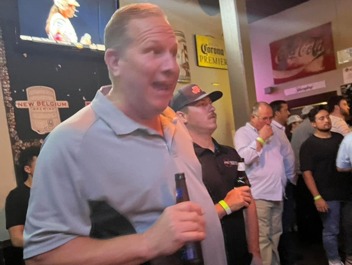 Man in a light blue shirt holding a beer at a bar, talking, surrounded by other people.