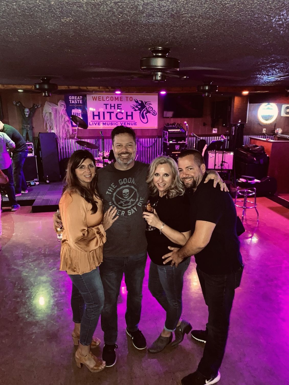 Four people posing together in a bar with a stage; two women, two men, all smiling.