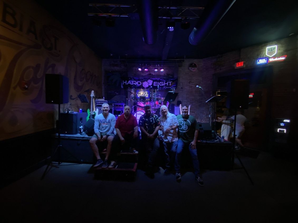 Band of five people seated on a stage, in front of instruments, under purple lighting.