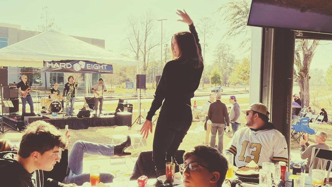 Woman dancing on a table near a band playing outdoors. People watch and eat. Sunny day.