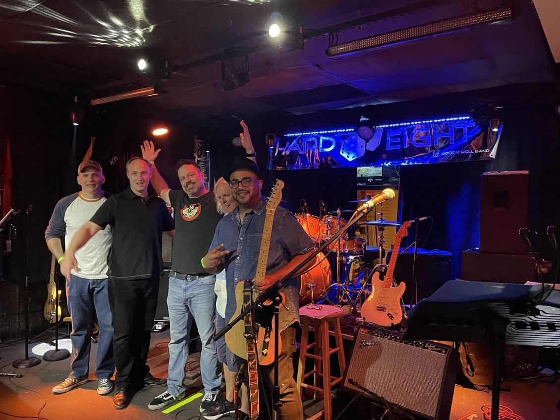 Band on stage, waving. Guitars, drums, and amplifiers. Dark venue with 