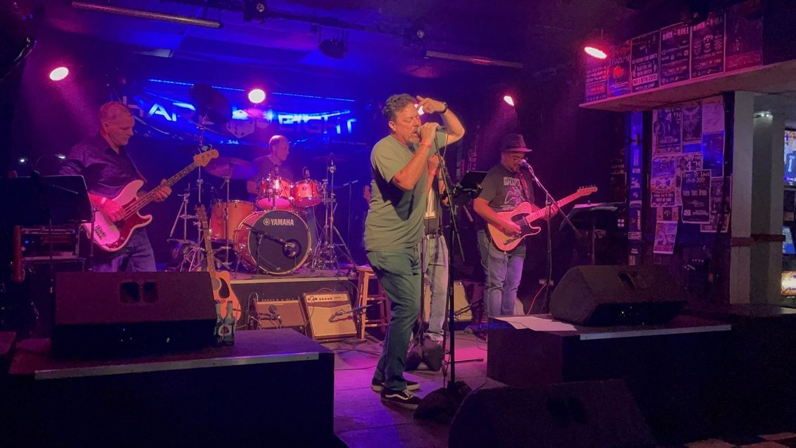 Band performing live on stage at a venue; singer with microphone, musicians playing instruments, stage lighting.