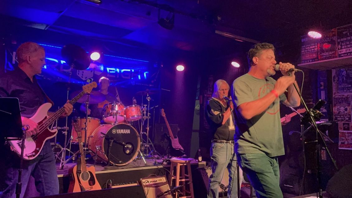 Band performing live on stage at a venue. Singer with microphone, bassist, drummer, other musicians; blue stage lighting.