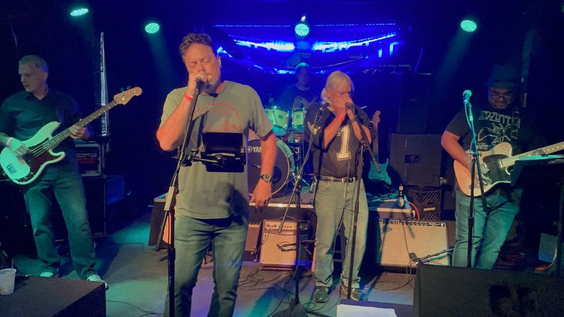 Band on stage: Bass guitar, lead vocal, harmonica, guitar. Blue stage lighting.