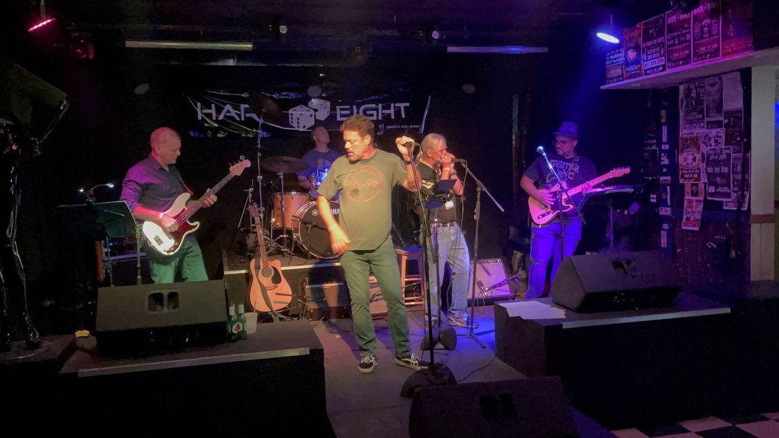 Band performing live on stage at a venue; musicians playing instruments, including violin, guitar, bass, and drums.