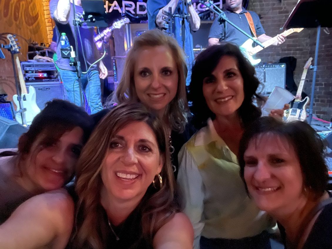 Five smiling women pose for a selfie in front of a band on a stage with brick walls.