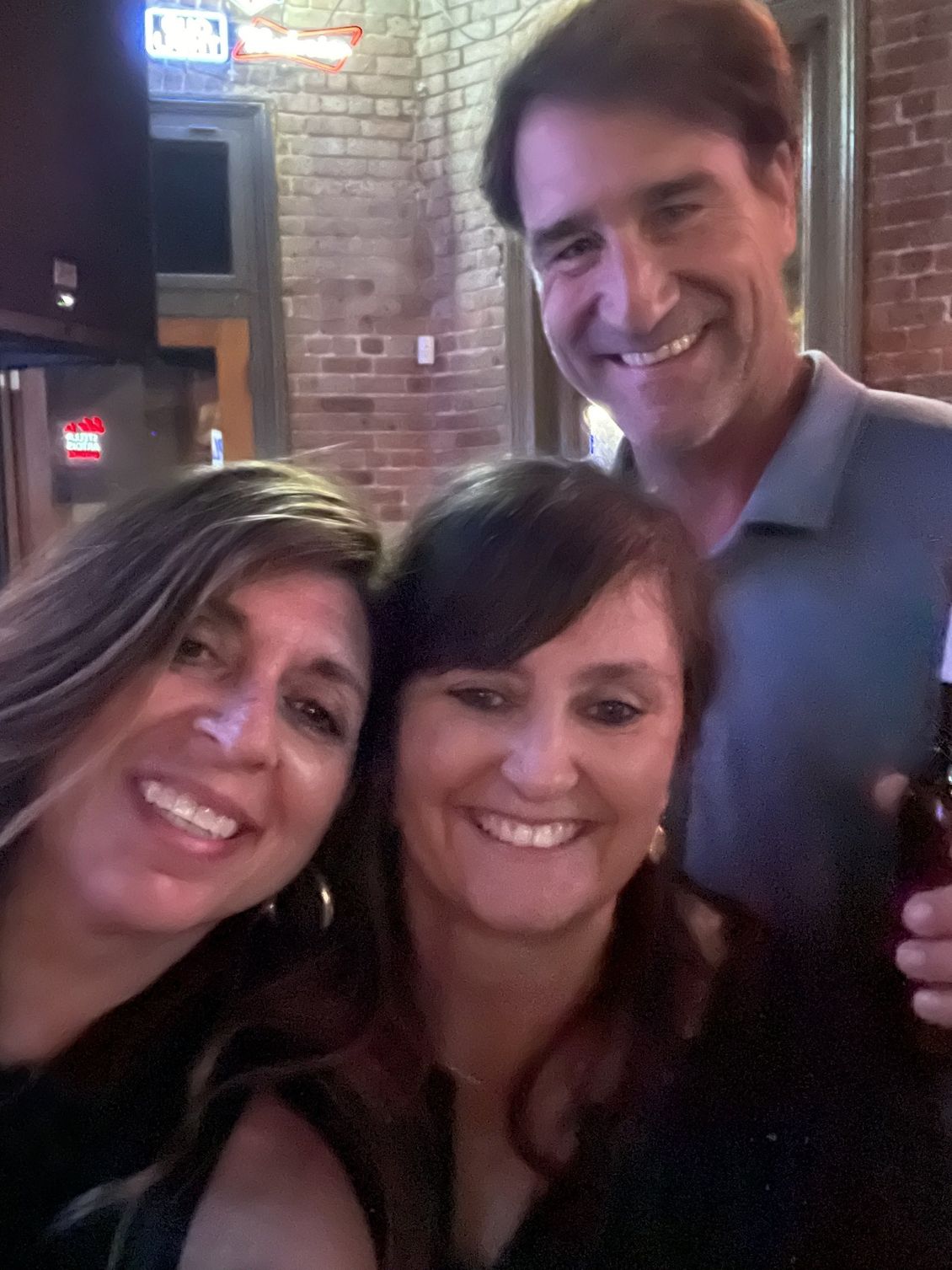 Three people smiling close together in a bar. Dark setting with brick wall, man holding bottle.