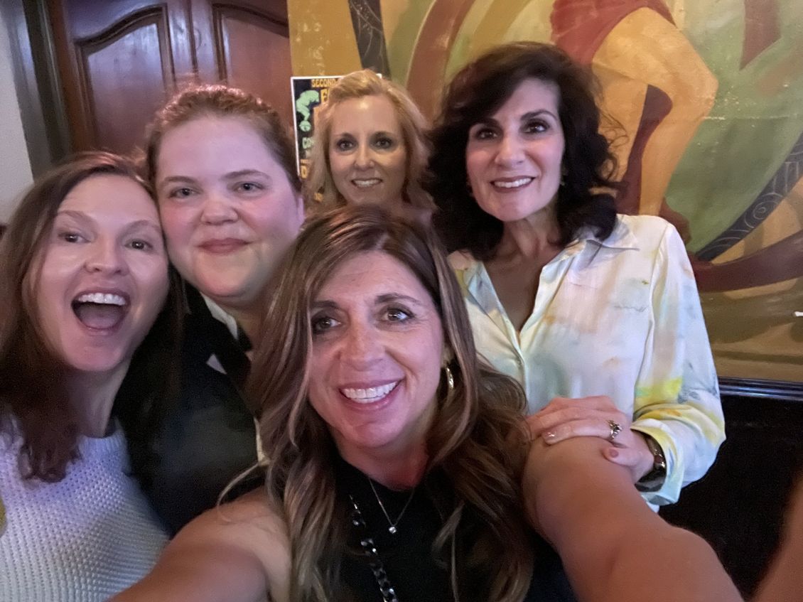 Five women smiling for a selfie; indoors with colorful artwork in the background.