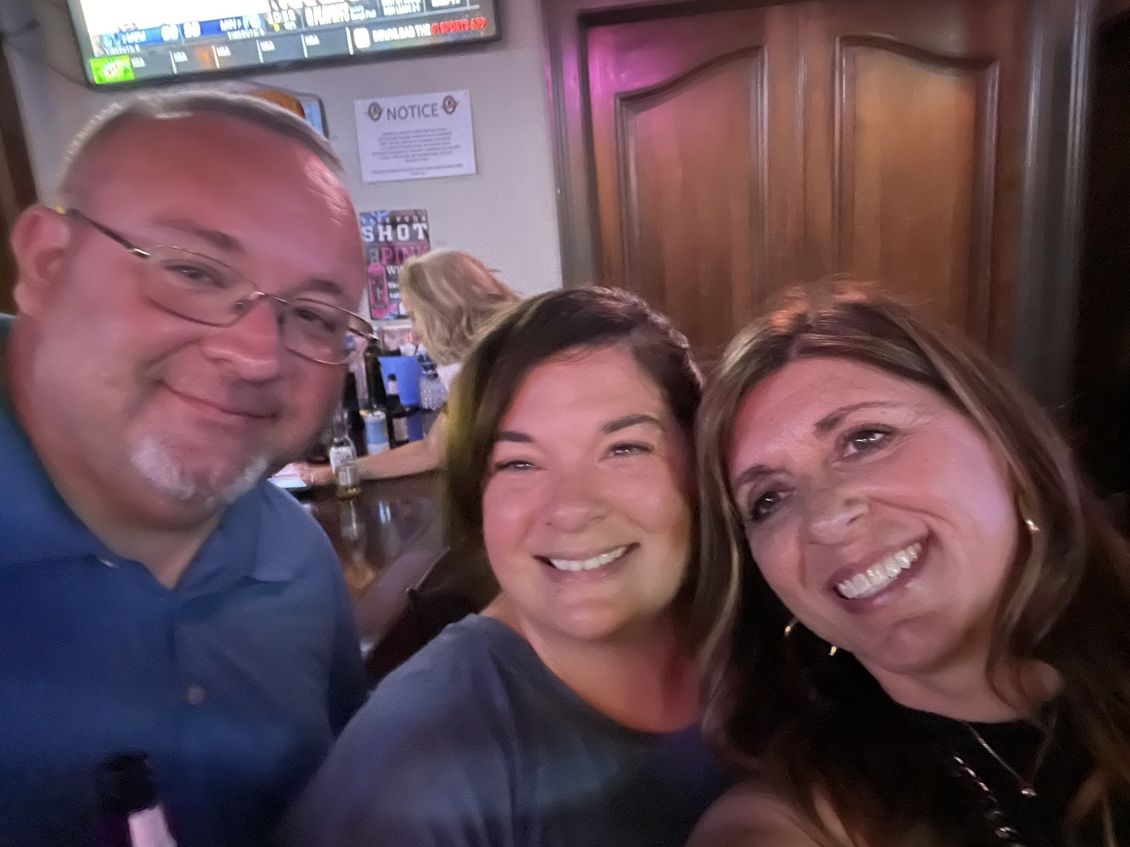 Three smiling people taking a selfie inside a bar. A bartender is visible in the background.