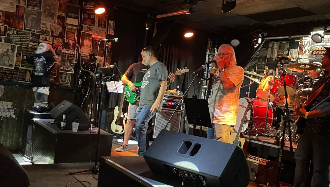 Band performing live on stage in a dimly lit bar. Instruments and crowd visible.