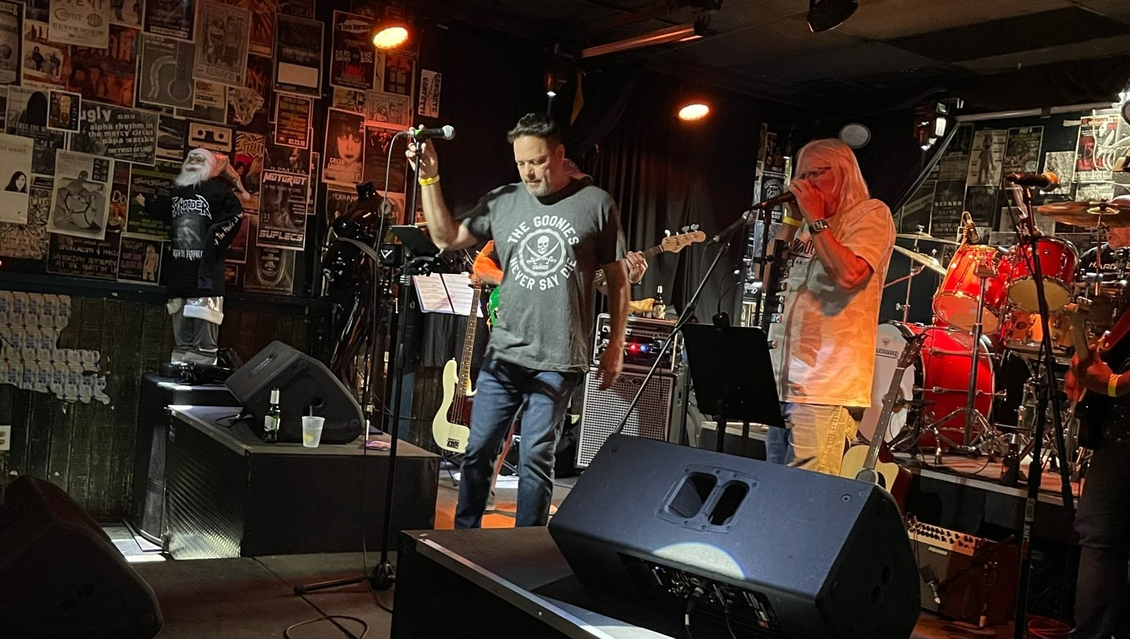 Two men on a stage with microphones, one holding up a drink. Music venue, band in background.