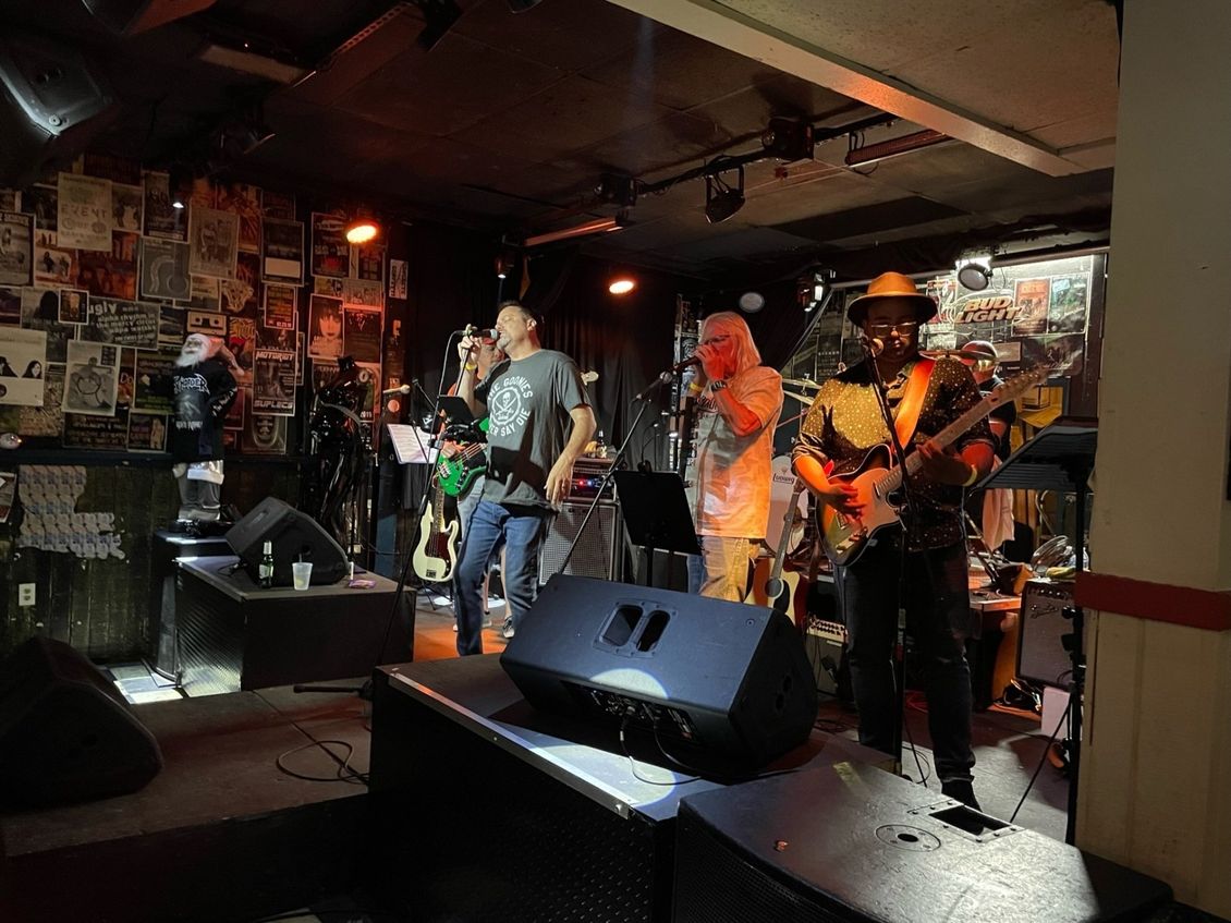 Band performing on stage in a dimly lit bar. Musicians with microphones, guitar, and lights. Posters on the wall.