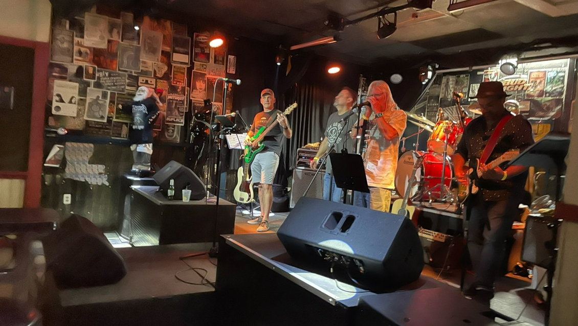 Band playing live on stage in a dimly lit club. Musicians with instruments in front of a wall with posters.