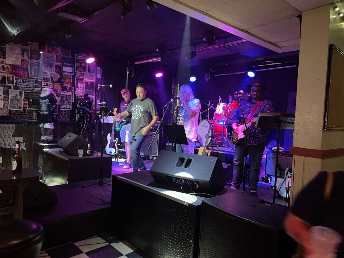 Band performing on a stage lit with purple and white lights in a bar.