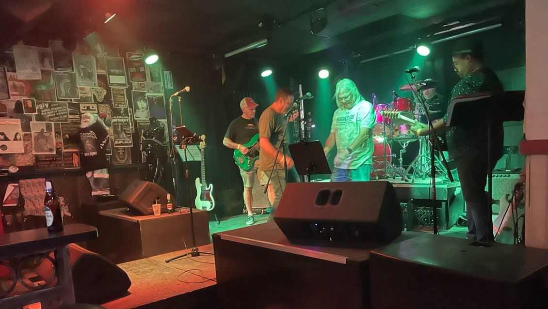 Band performing on stage in a dimly lit venue with green stage lighting. Several musicians play instruments.