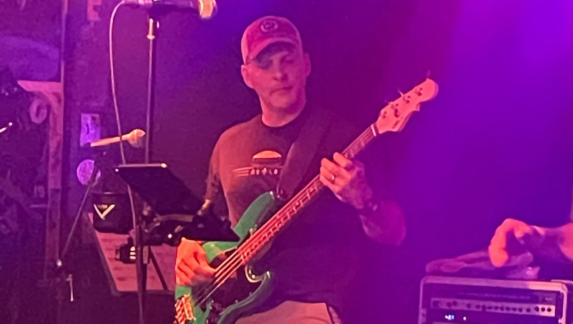 Man playing a green bass guitar on stage, wearing a cap and dark shirt. Purple and white lighting.