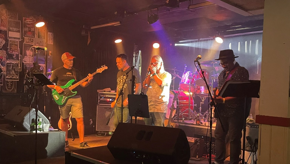 Band performing on stage under stage lights. Musicians playing instruments, singing into microphones. Dim setting.