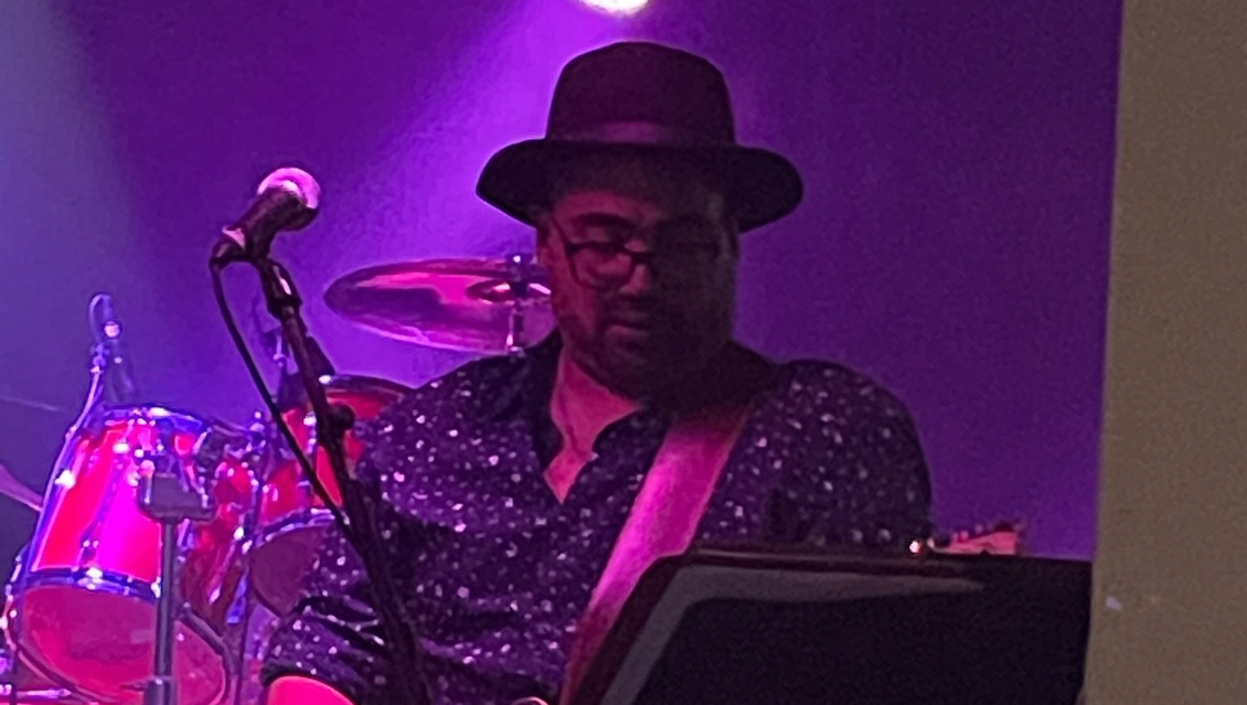 Keyboard player with a fedora, performing on stage with a microphone and drum set, bathed in purple light.