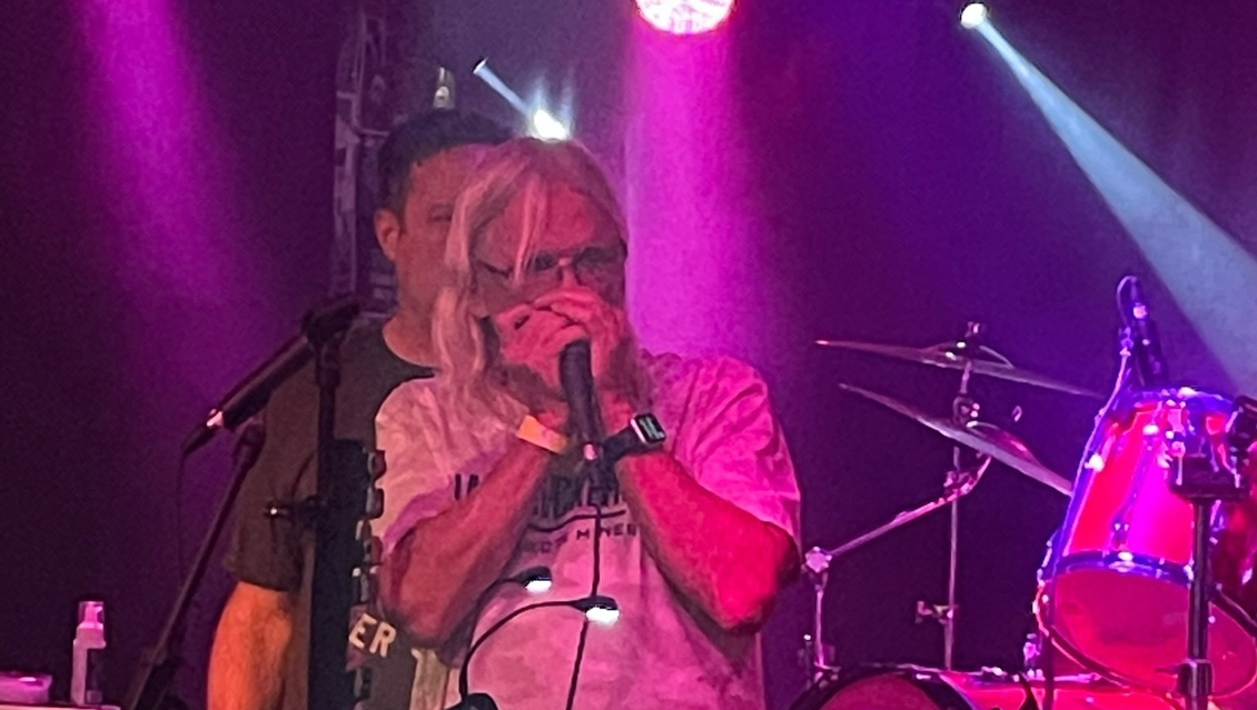 Man playing harmonica on stage, lit by pink and white lights, near a drummer.