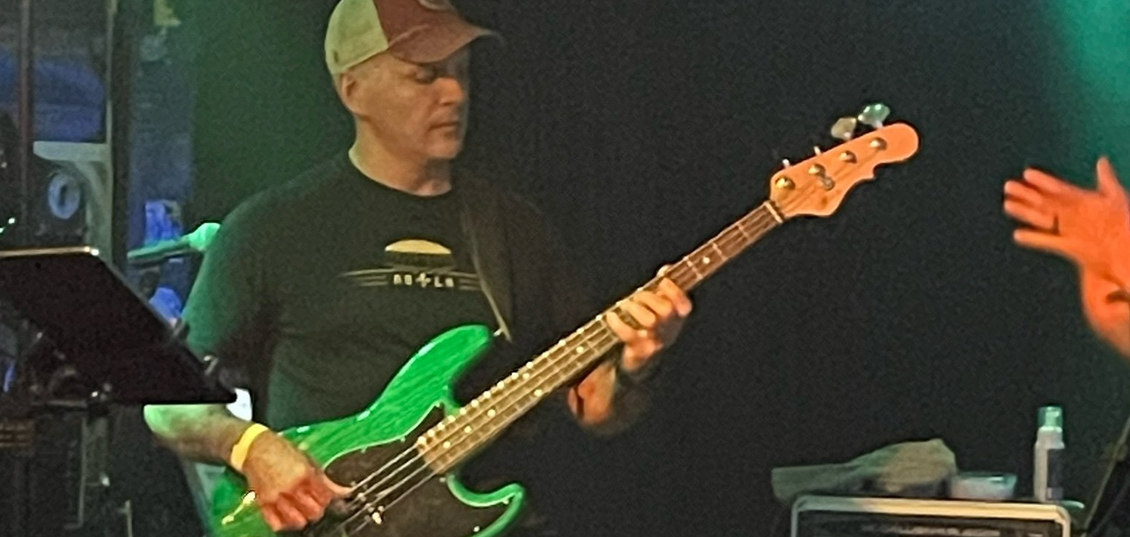 Man playing a green bass guitar on stage with a tan hat and black t-shirt.
