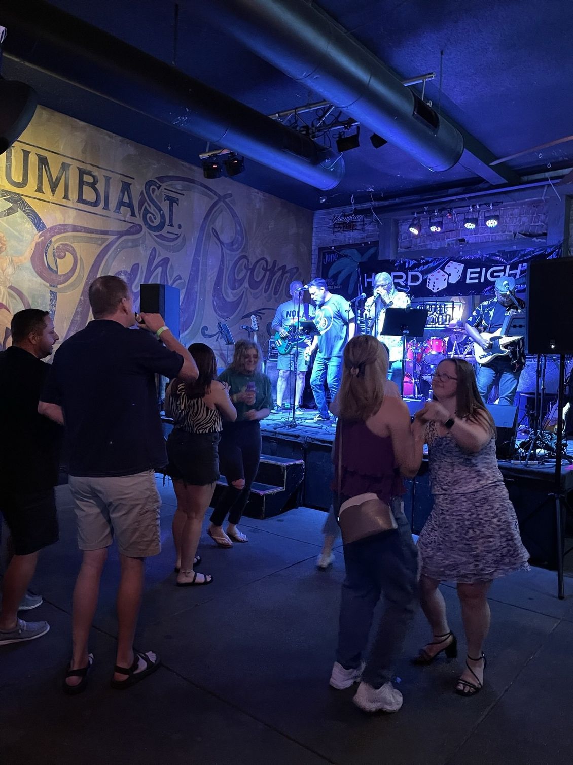 People dancing in a bar while a band performs on stage with blue lighting.
