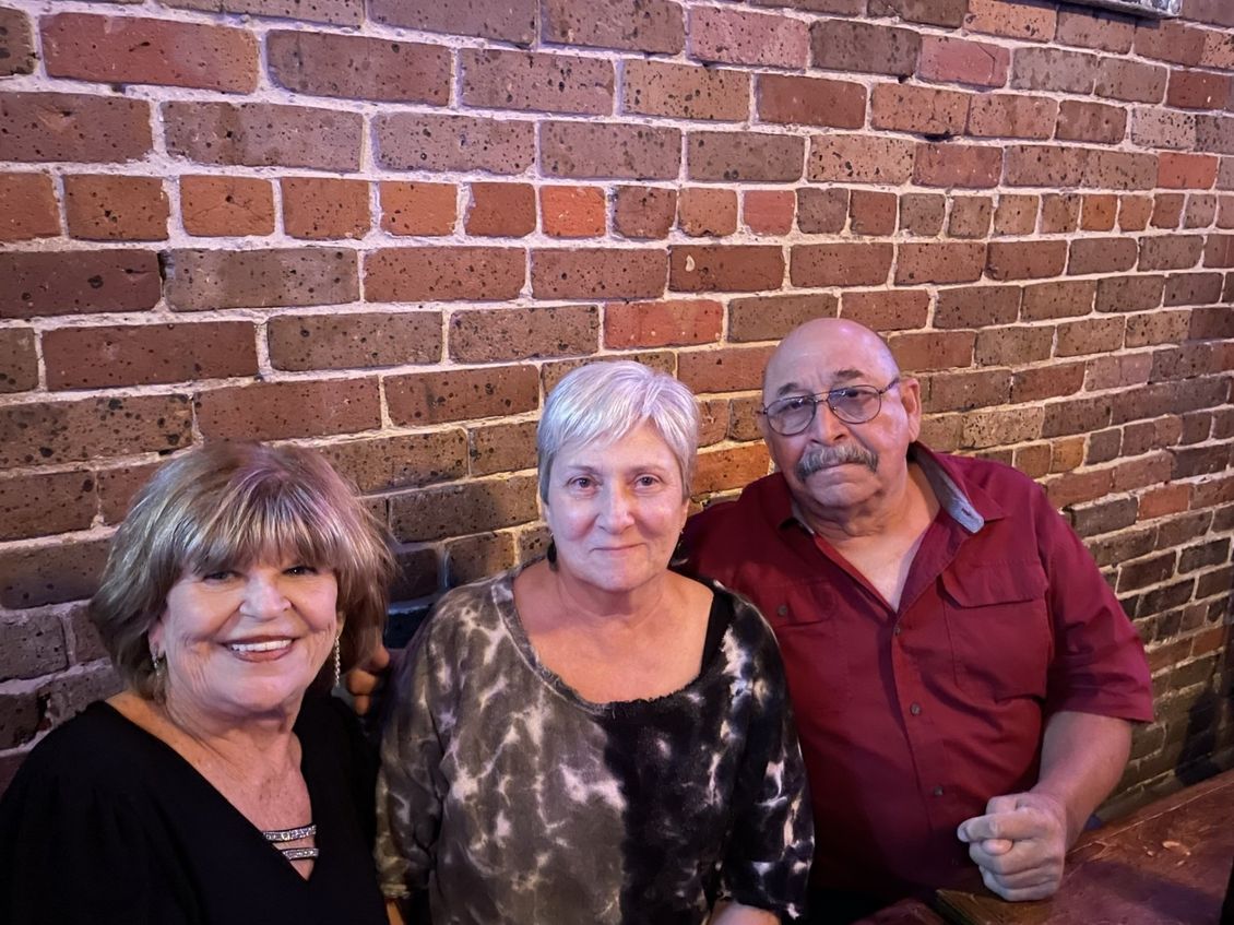 Three people pose in front of a brick wall. Woman in black top smiles, two others face forward.