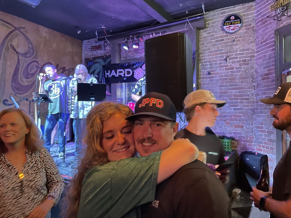 Couple embracing in front of a band on a stage in a bar; people watching, brick wall, and purple lighting.