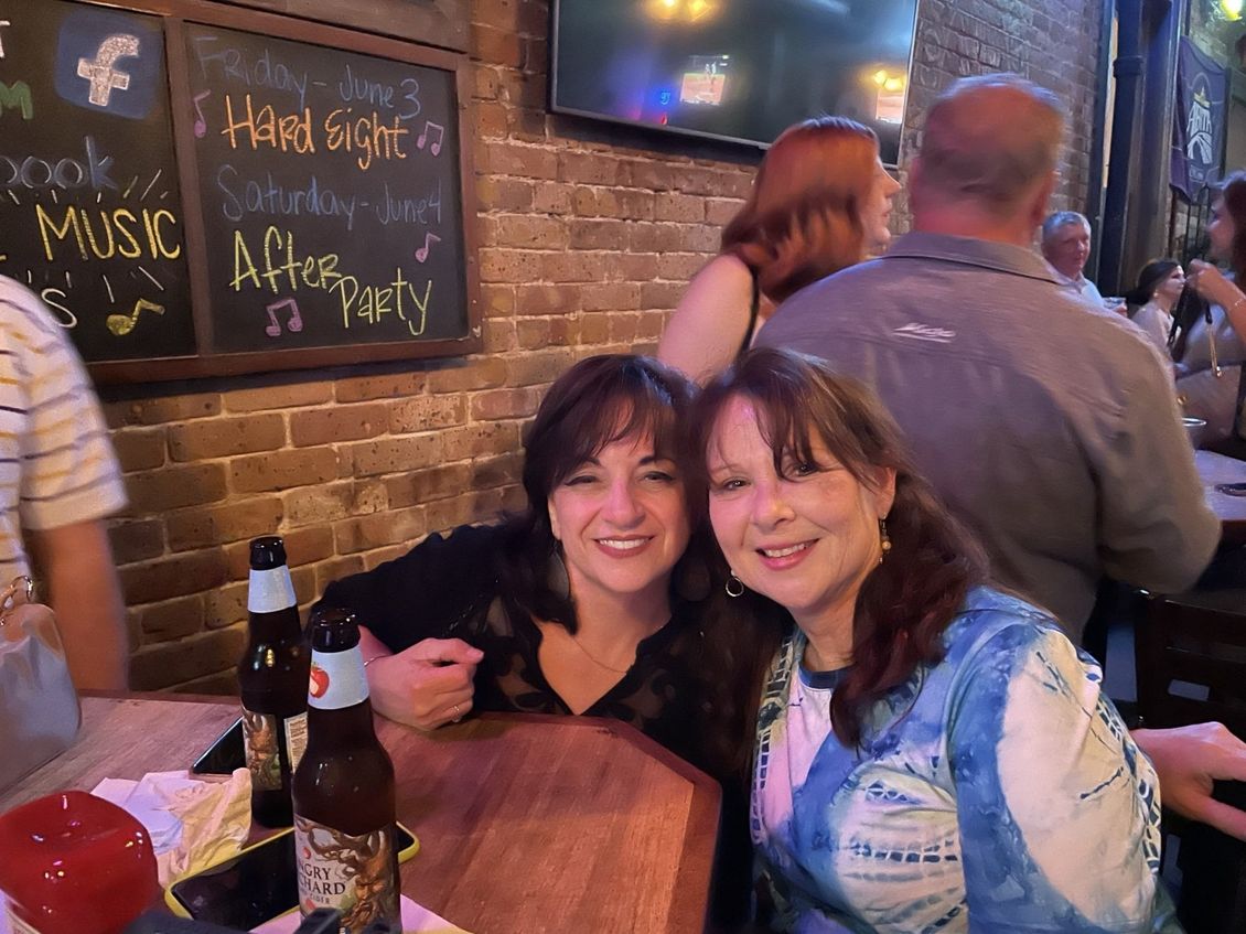 Two women smiling at a bar, leaning on a table with beer bottles. A chalkboard sign is visible.
