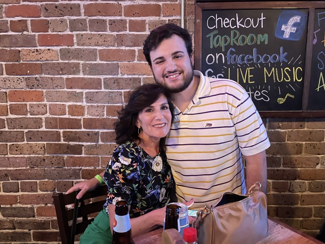 Woman and man smiling, embracing, near a brick wall. Man wears striped shirt, woman floral top.