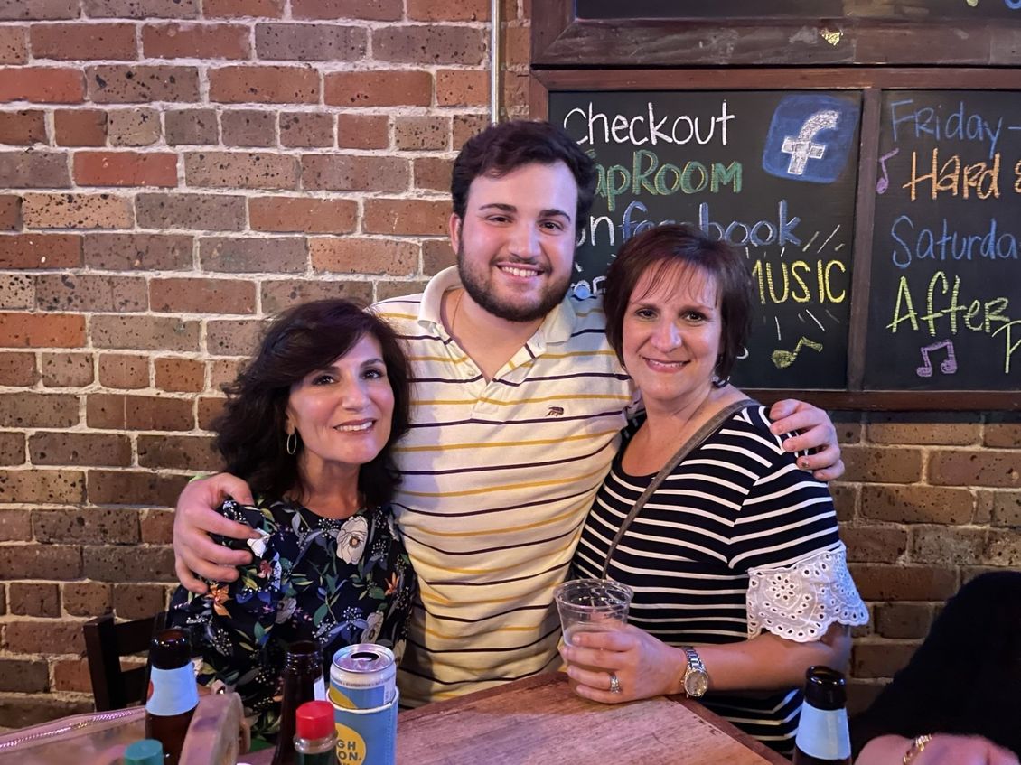 Three people smiling and posing in front of a brick wall, possibly a bar or restaurant. One man has his arms around two women.