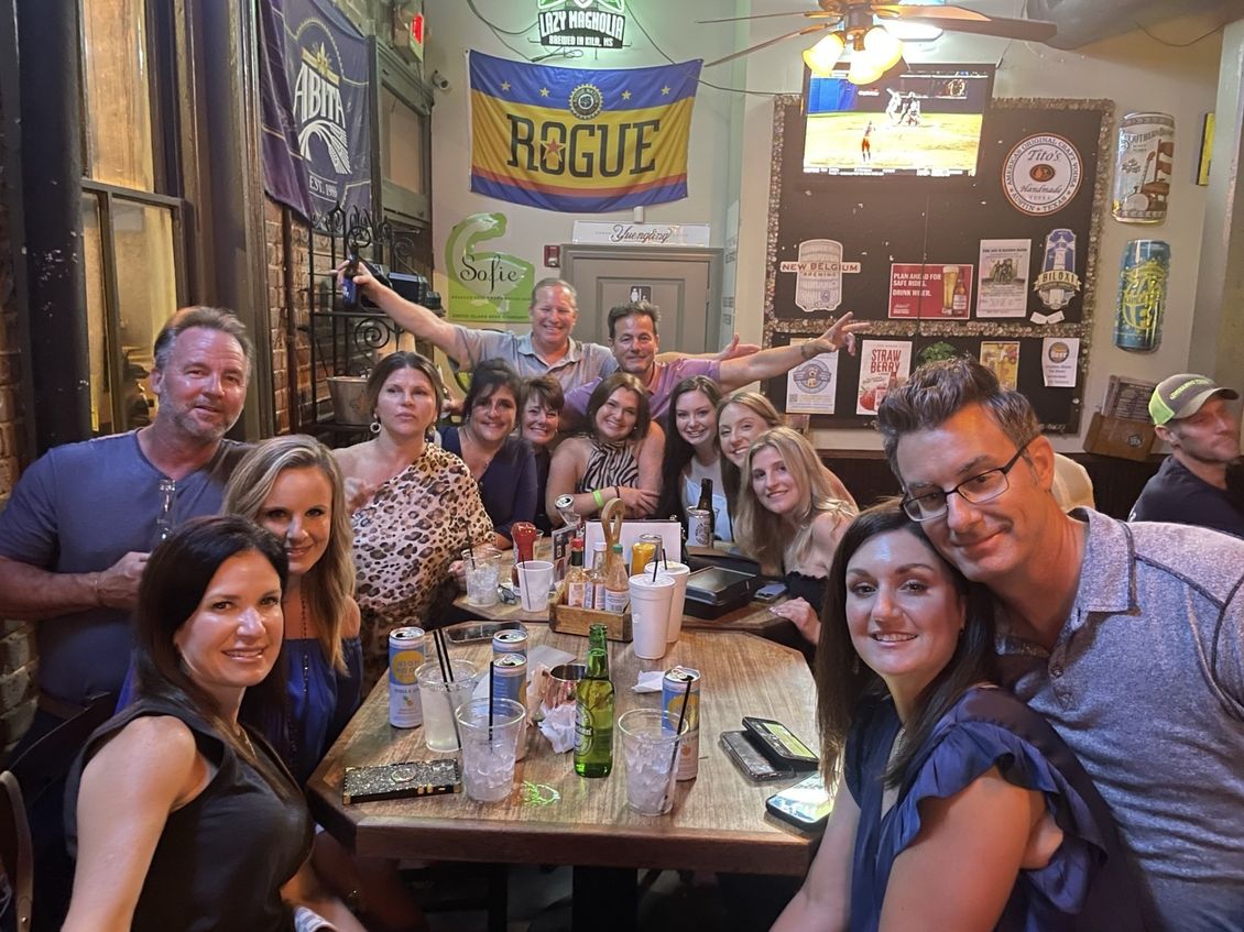 Group of people smiling at a table in a bar; drinks, Rogue banner, TV screen in background.