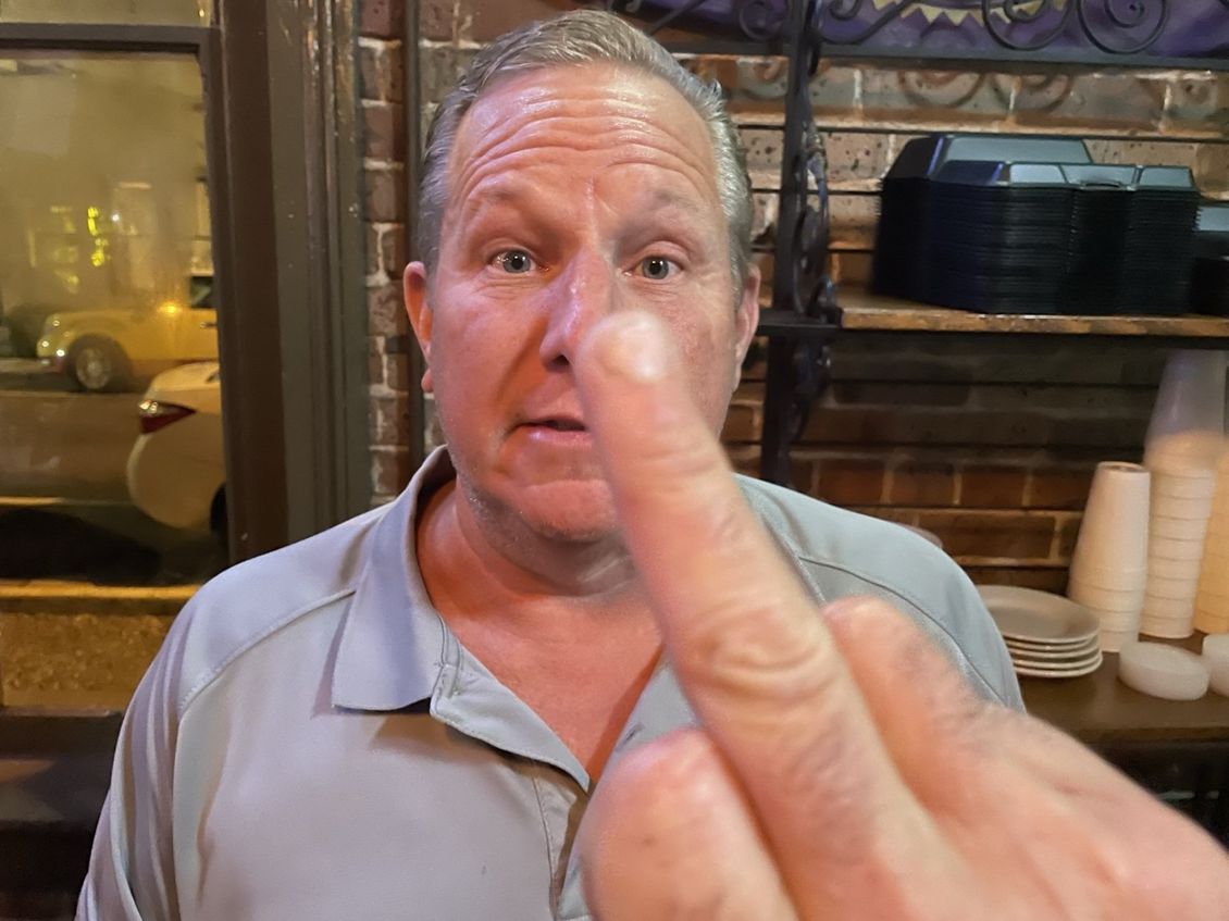 Man giving the middle finger, standing outside a brick building.