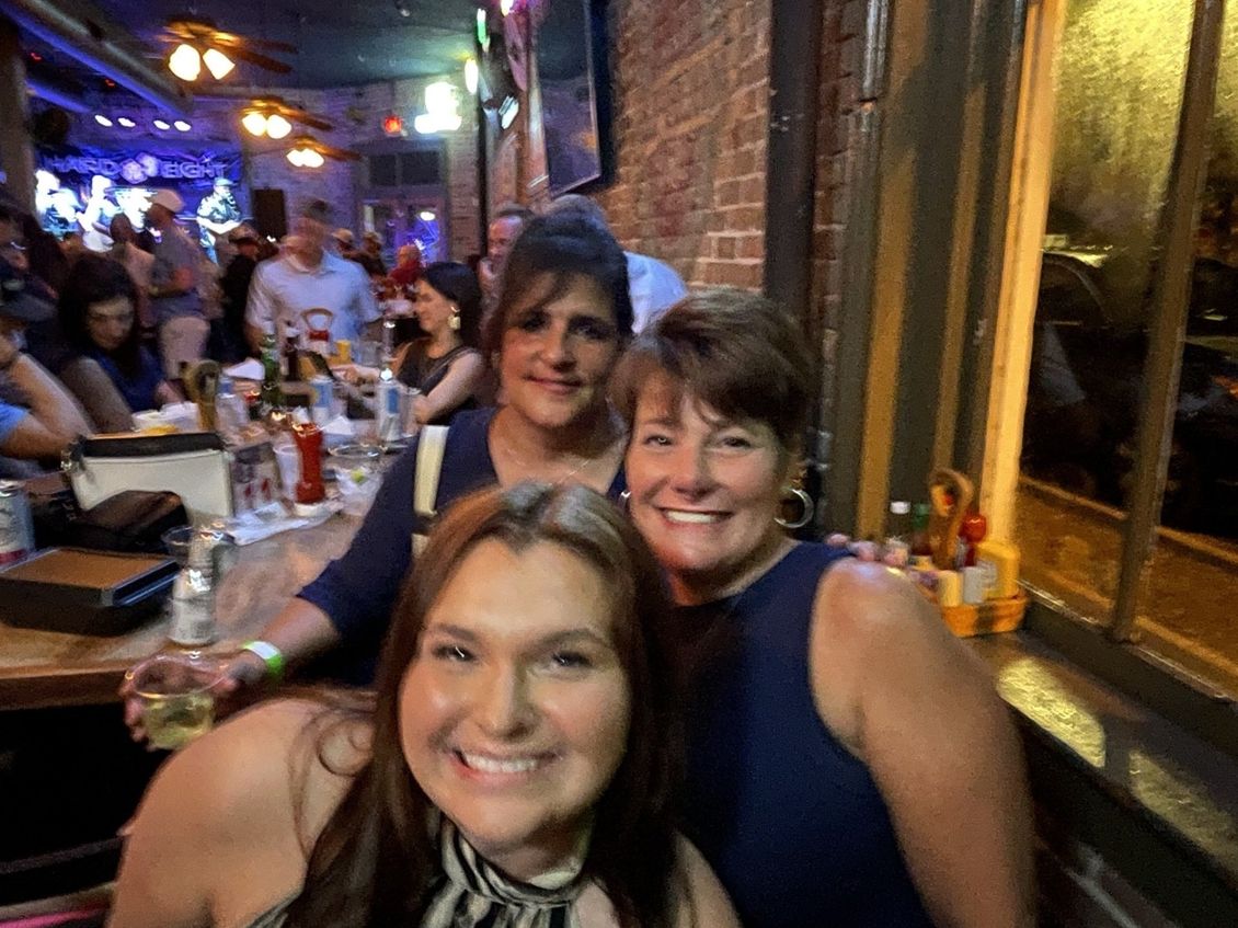 Three smiling women pose for a selfie in a dimly lit bar, brick wall background.