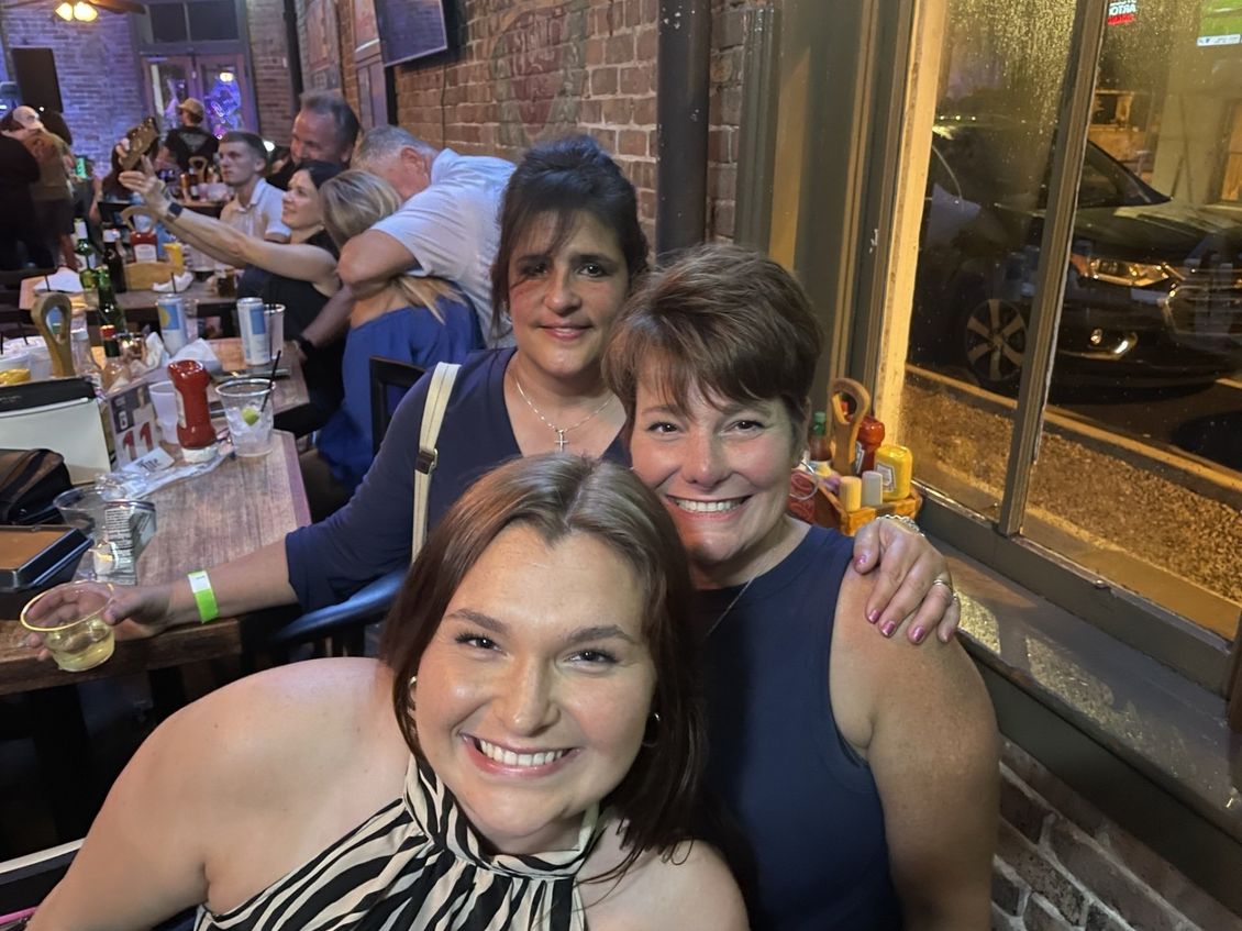 Three smiling women pose for a photo in a bar; one in front, two behind. Brick wall and window visible.