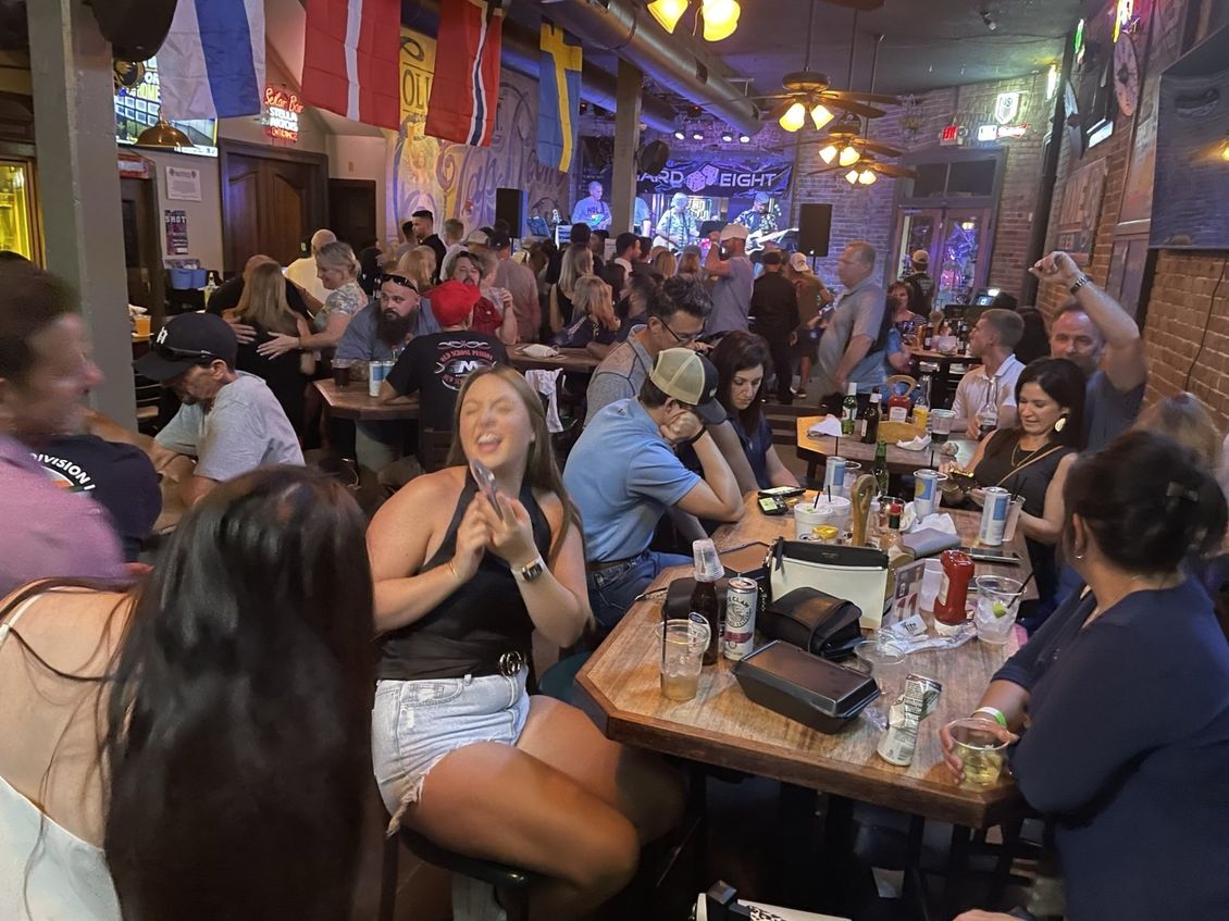 A crowded bar with a live band playing. People are sitting at tables, laughing, and enjoying drinks.