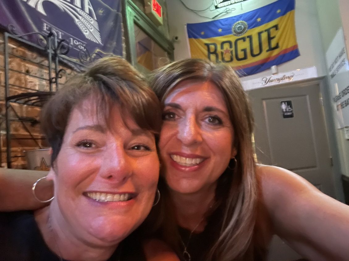 Two smiling women take a selfie in a bar, Rogue flag in the background.