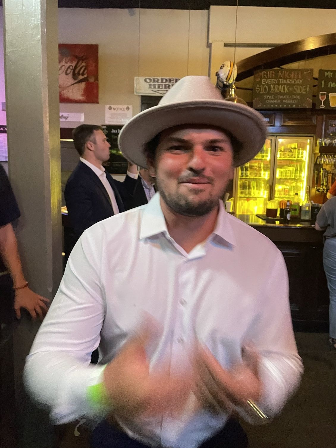 Man wearing a hat and white shirt smiles, indoors at a bar, clapping hands.