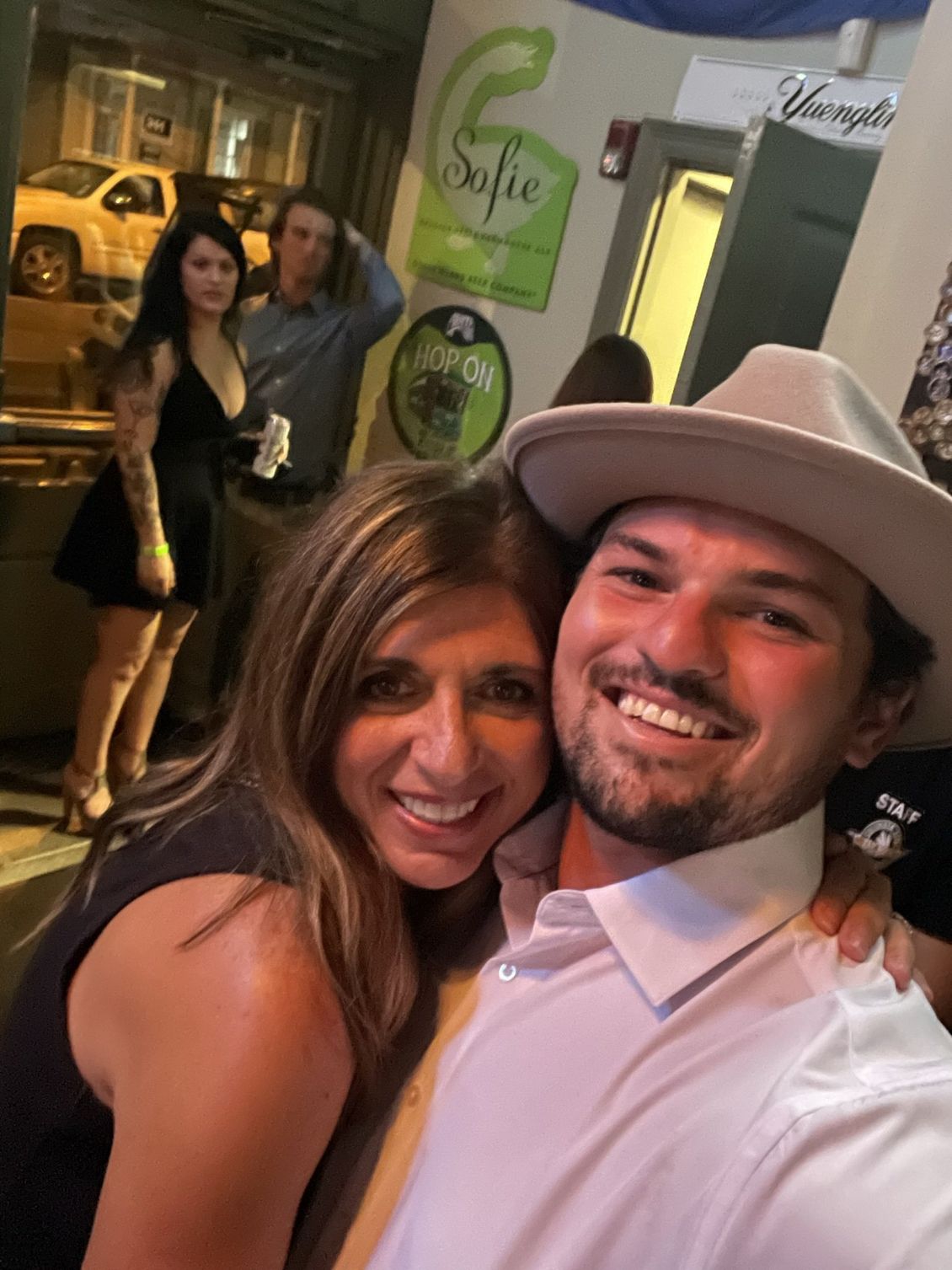 Two people smiling, hugging, near bar entrance; others visible in background.