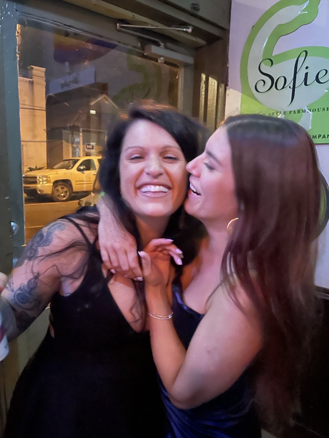 Two women embrace and smile, one whispering in the other's ear, near a door.